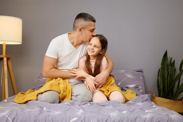 A father lovingly embraces his daughter on a bed draped with blankets in a softly lit room. They...