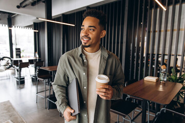 Smiling freelancer holding laptop and coffee walking in coworking space