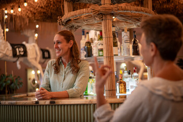 Happy woman smile stands behind a bar counter in a tropical or rustic-themed includes a few plants, enhancing the natural and cozy atmosphere holidays concept.