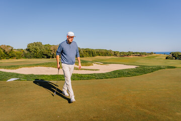 A golfer walking on a sunny golf course with a club in hand, sand bunker, green fairway, and clear blue sky in the background.