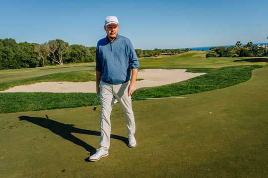 A golfer walking on a putting green with a golf club, sand bunker, and lush fairway in the background under a sunny blue sky.