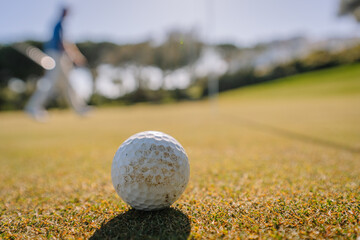 Close-up of a golf ball on a grassy course with a blurred golfer in motion in the background under bright sunlight and a clear sky.