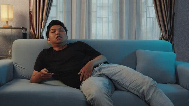 Young man with a dull expression watching TV on the sofa. Attempting to distract himself from feelings of boredom and loneliness. TV viewing, home boredom, young male, sofa relaxation, solitude