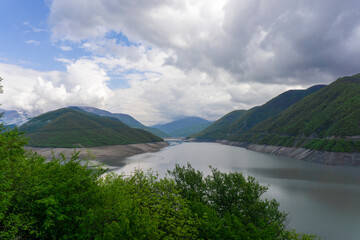 Reservoir on the river between two hills. Steep rocky banks. Spring green grass, bushes, trees. Mountains, hills and cloudy sky in the background