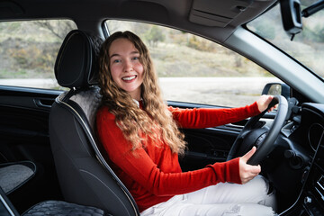 Naklejka premium A young friendly positive woman is holding a car steering wheel, looking at the camera and smiling. Portrait of a cheerful female driver in the driver's seat wear a casual red jumper. Lifestyle.