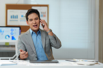 Young businessman sitting at his office desk, talking on a mobile phone while arguing with someone, gesturing expressively with his hands, displaying signs of stress and frustration