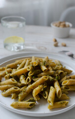 Penne pasta with pistachio pesto with a sprig of basil in a white plate with a glass of water with lemon on a table in a restaurant. Italian cuisine concept. Vertical orientation. Selective focus.