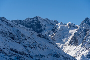 Obraz premium Dombay. Karachay-Cherkessia. Giant rocks with snow-capped peaks and glaciers rise majestically against blue autumn sky. Pine and spruce forests grow at foot of mountains. Sunny day. Autumn 2024