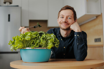 Young smiling man vegan eating a green salad in big bowl at kitchen