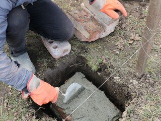 worker in orange gloves laying and leveling a layer of fresh cement with a trowel in a hole in the ground on a block with a string stretched nearby as a level, preparing for foundation work