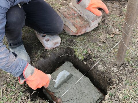 worker in orange gloves laying and leveling a layer of fresh cement with a trowel in a hole in the ground on a block with a string stretched nearby as a level, preparing for foundation work