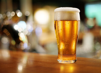 Glass of chilled beer on wooden bar table top and blurred bar interior at the background.