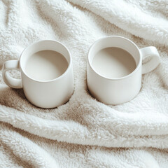 Two white mugs filled with warm beverage rest on soft, fluffy blanket, creating cozy and inviting atmosphere. top down view highlights simplicity and warmth of scene
