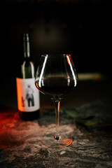 Elegant red wine glass next to bottle on dark surface with subtle reflections and warm light effects