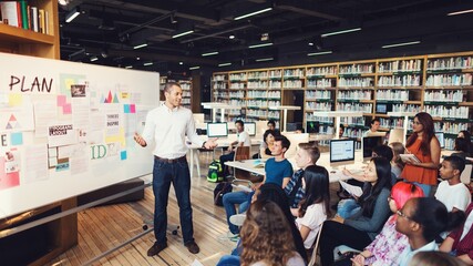 Diverse group of students in a library listens to teacher presenting at a whiteboard. Diverse students study in the library with teacher teaching in front of whiteboard. Students join class at library