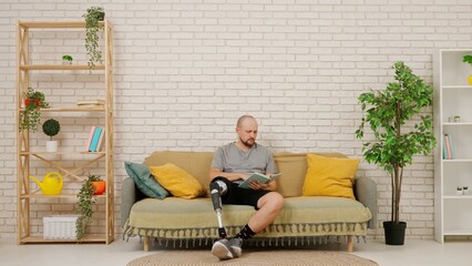 A man with a prosthetic leg reads a book sitting on a couch in a room with a modern home interior © kinomaster