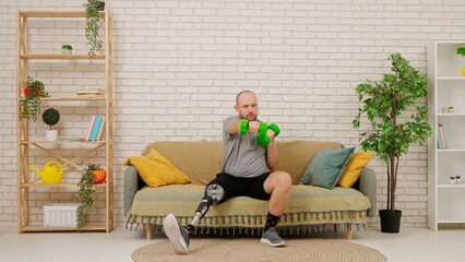 A man with a prosthetic leg sits on a couch in a cozy living room, doing an exercise with dumbbells, training his arms.