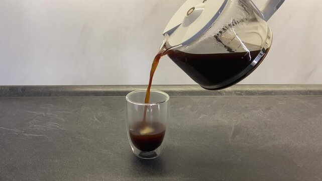 Black coffee is poured from a coffee pot into a transparent glass cup.