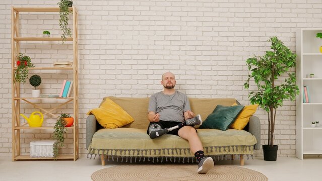A man with a prosthetic leg sits on a couch in a cozy living room and meditates.