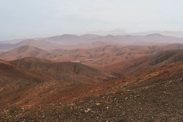 Gordijnen A barren, rocky mountain landscape in Fuerteventura under a cloudy sky. The mountains are brown and textured, with sparse vegetation in the foreground © Rokas