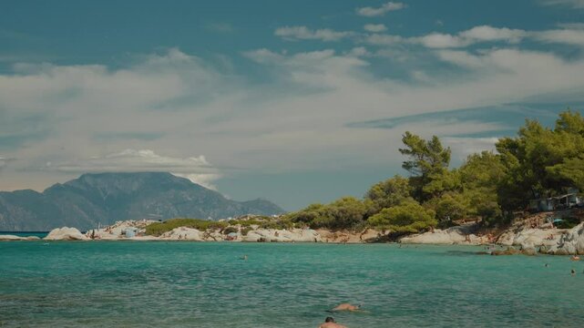 Tourists sunbathing and swimming, enjoying clear turquoise waters, along the picturesque Greek coastal landscape under bright summer skies