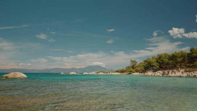 Tourists enjoying crystal clear turquoise waters, sunbathing and swimming along picturesque Greek coastal landscape under bright summer sky