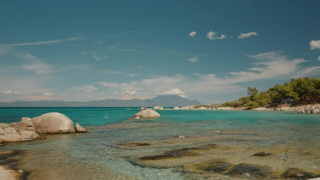 Mediterranean Sea coast with tourists enjoying crystal clear turquoise waters, sunbathing and swimming along picturesque Greek coastal landscape under bright summer sky