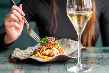 A close-up of a woman’s hand holding a fork, ready to eat a gourmet dish with fresh ingredients, accompanied by a glass of white wine on an elegant table setting.