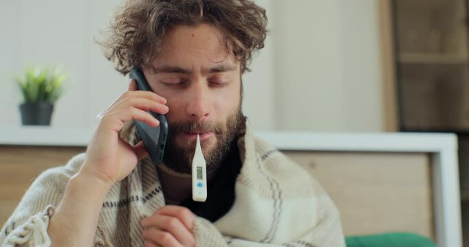 Sick young man with termometr in mouth talking on the phone while sitting on the sofa, sneezing and caughing as having a cold. Concept of healthcare and illness.