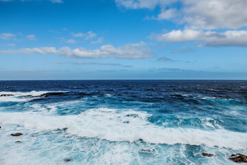 Dramatic ocean waves crashing against the rugged volcanic coastline of El Hierro, Spain. The deep blue sea contrasts with foamy white water, evoking power and nature’s force...