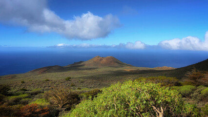 Scenic volcanic landscape overlooking the ocean with lush green vegetation. The contrast of blue sky, clouds, and rugged terrain highlights the beauty of nature.