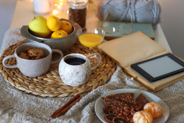 Books, e-reader, tea, orange juice, fruit, snacks, yarn, blanket and lit candles on the table. Autumn or winter hygge at home. Selective focus.