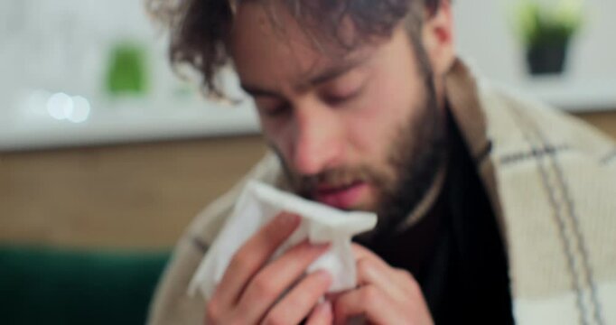 Close up of the young sick man in the warm scarf and a plaid blowing his nose in the napkin as he having a cold.