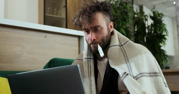 Close up ill male sitting on sofa at home office under plaid, working remotely, using paper tissue with termometr in mouth. Man feeling unwell, sick with running nose, need a rest.