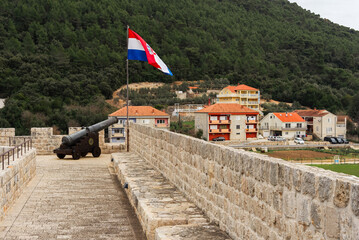 Historical cannon in the town of Ston, Croatia. A flag flies above the cannon. In the background are modern homes.