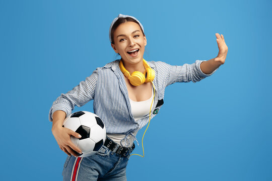Energic young woman holding soccer ball and headphones dance over blue background