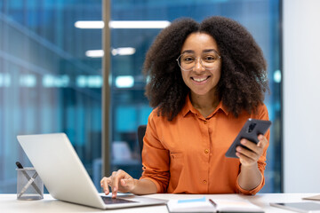 Portrait of a young successful woman at her workplace with a laptop and a smartphone in her hands....