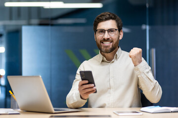 Portrait of a happy young man who is in the office, sitting at a desk with a laptop, holding a mobile phone and rejoicing in success shows a yes and victory gesture, looks satisfied at the camera