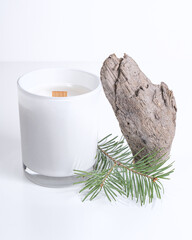 White candle in a wooden wick on a white background, next to a tree and a spruce branch, symbolizing the aroma of a candle