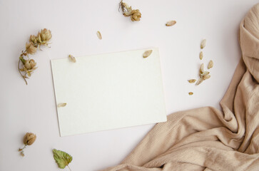 Blank paper card on white background with dried plants and wavy beige fabric, top view. Boho mock-up scene with greeting card template