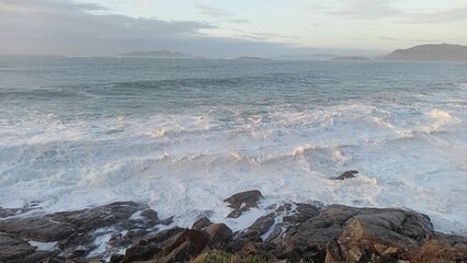 Obraz premium Stunning coastal scene featuring powerful ocean waves crashing against rugged rocks under a serene sky, with distant islands on the horizon. Perfect for nature, travel, and seascape themes