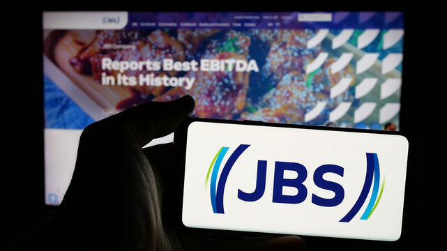 Stuttgart, Germany - 01-30-2025: Person holding mobile phone with logo of Brazilian meat processing company JBS S.A. on screen in front of business web page. Focus on phone display.