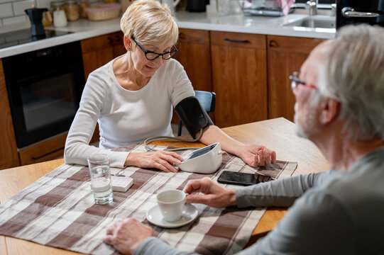 An elderly woman checks her blood pressure at home using a digital monitor while an elderly man sits across from her at the kitchen table with a cup of coffee - Powered by Adobe