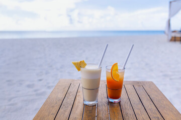 Two cocktails with slices of fruit are on a wooden table on the beach. Delicious chilled drinks on the background of tropical trees and the ocean.