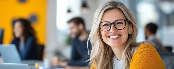 A joyful young woman with glasses smiles at the camera in a modern office space, showcasing a vibrant and collaborative work environment filled with engaged colleagues.