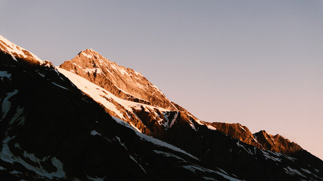 Vue  a&eacute;rienne d'un sommet montagneux, partiellement recouvert de neige, illumin&eacute; par la chaude lumi&egrave;re dor&eacute;e du soleil levant. Paysages alpins vus par drone. 