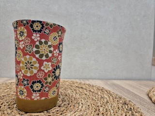 Red cup with floral pattern and golden bottom standing on round wicker placemat on wooden table with gray wall background, representing a stylish and cozy dining or tea time setting