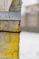 This photograph captures a close-up of a weathered metal pole with peeling yellow paint and a rusted wire wrapped around it. The background shows a blurred winter scene with snow-covered ground 