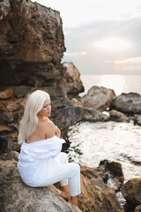 Attractive young adult woman 30-35 year old with long blonde hair wearing white shirt and denim pants sitting on rocks over sea at sunset. Summer vacation season. 