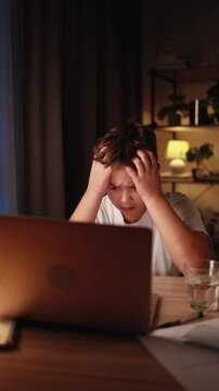 Stressed young boy holding head in frustration sitting at desk in cozy evening room emotionally reacting to laptop screen overwhelmed by online task game or remote schoolwork